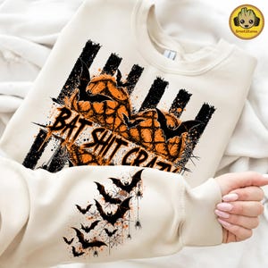 May include: A cream-colored sweatshirt featuring a graphic print of orange pumpkins, black bats, and spiderwebs. The text "BAT SHIT CRAZY" is printed across the center. The sleeve displays a cascading pattern of black bats and spiderwebs.