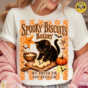 May include: A white t-shirt featuring a graphic of a black kitten on a wooden cutting board with a cookie, a bowl of flour, and a pumpkin. The text reads "SPOOKY BISCUITS BAKERY" and "WE KNEAD EM YOU NEAD EM" in orange and brown lettering.