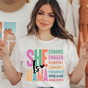 K&ouml;nnte beinhalten: Ein wei&szlig;es T-Shirt mit einem bunten Grafikdesign, auf dem "She is Mama" steht, darunter aufgelistete Bibelverse. Die Verse sind: Phil 4:13 Strong, 1 Thess 1:4 Chosen,  Ezek 16:11 Beautiful,  Mark 10:27 Capable,  Rom 8:37 Victorious,  Deut 31:6 Never Alone,  1 John 4:19 Greatly Loved.