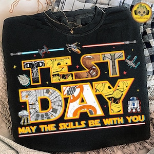 Peut inclure: Sweat-shirt noir avec l'inscription "TEST DAY" en jaune et blanc, représentant des personnages et éléments de Star Wars. La phrase "MAY THE SKILLS BE WITH YOU" est imprimée en dessous. Comprend des sabres laser et des vaisseaux spatiaux.