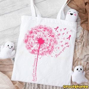 May include: White tote bag featuring a pink dandelion design with the words "Breast Cancer Awareness" and "Never give up." Pink ribbons are scattered around the dandelion. The bag has two white handles.