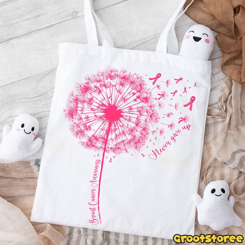 May include: White tote bag featuring a pink dandelion design with the words "Breast Cancer Awareness" and "Never give up." Pink ribbons are scattered around the dandelion. The bag has two white handles.