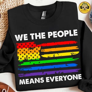 May include: A black long-sleeve sweatshirt featuring the white text WE THE PEOPLE at the top and MEANS EVERYONE at the bottom. Between the text is a distressed graphic of a flag with horizontal rainbow stripes and black stars on a yellow field.