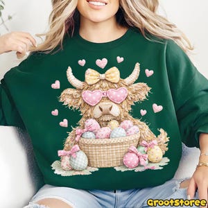 Puede incluir: Sudadera verde oscuro con un dise&ntilde;o de vaca de las Tierras Altas. La vaca lleva gafas de sol en forma de coraz&oacute;n y un lazo, y sostiene una cesta de huevos de Pascua. Hay corazones rosas dispersos. El texto "GrootStoree" est&aacute; abajo.