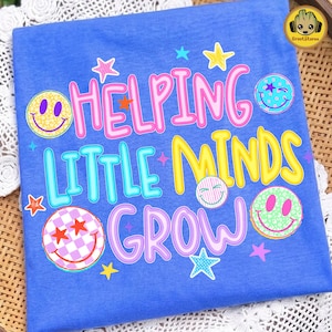 May include: A blue t-shirt featuring the text HELPING LITTLE MINDS GROW in pink, blue, yellow, and purple lettering. The design includes various colorful smiley faces with patterns like leopard print, checkers, and polka dots, along with small stars.