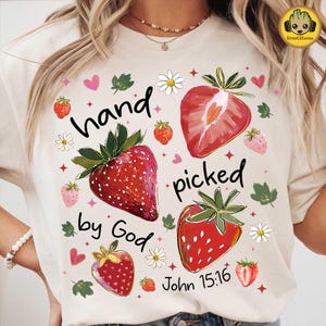 Puede incluir: Camiseta color crema con un diseño de fresas y el texto "hand picked by God John 15:16". El diseño incluye fresas rojas, corazones rosas, margaritas blancas y hojas verdes.