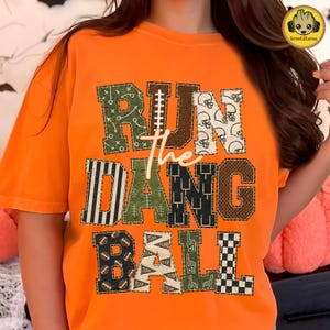 May include: An orange short-sleeve t-shirt featuring the text "RUN THE DANG BALL" in large, block-style letters. Each letter is filled with various football-themed patterns, including helmets, footballs, stripes, and play diagrams in green, brown, and white.