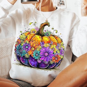 May include: White sweatshirt featuring a vibrant, watercolor-style pumpkin design. The pumpkin is adorned with colorful flowers in shades of purple, orange, blue, and green. The design has a whimsical, artistic aesthetic, perfect for fall.