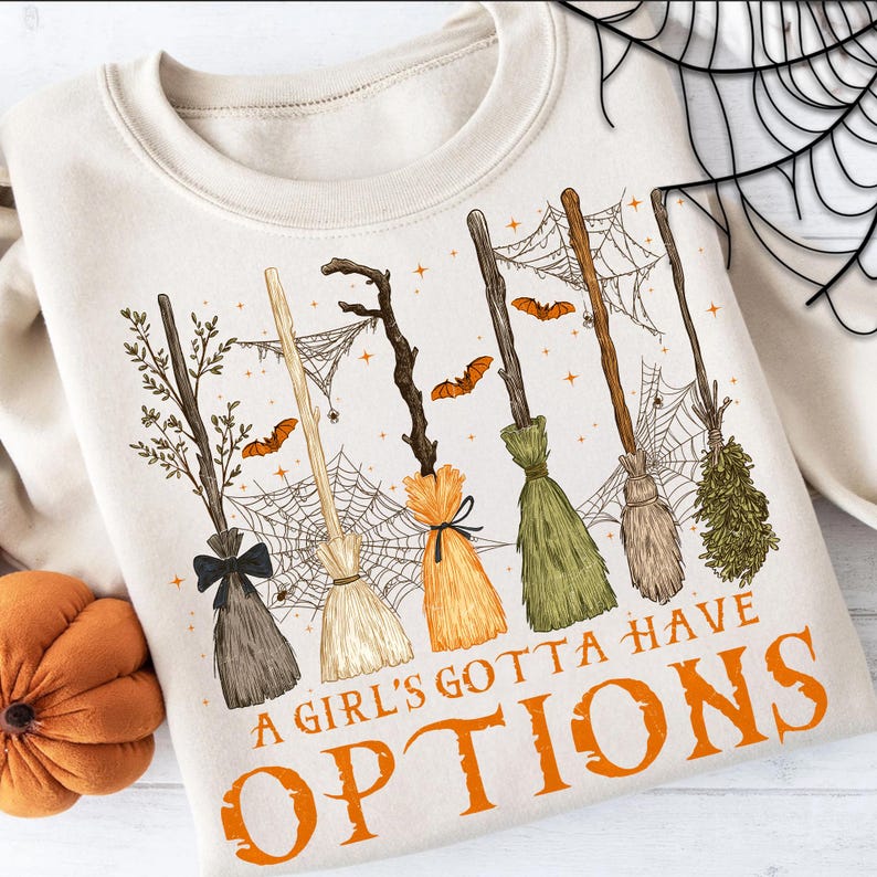 May include: Beige sweatshirt with a Halloween-themed graphic. The design features six different broomsticks, spiderwebs, and bats. The text "A Girl's Gotta Have Options" is printed in orange. A pumpkin is visible in the lower left corner.