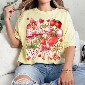 May include: A yellow t-shirt with a graphic print of strawberries, bows, and daisies. The strawberries are red with green leaves and the bows are pink with gold accents.