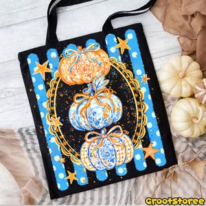 Puede incluir: Un bolso tote negro con un dise&ntilde;o vertical de tres calabazas apiladas con patrones florales en naranja y azul. Las calabazas est&aacute;n encerradas en un marco ovalado dorado rodeado de rayas azules, puntos blancos y peque&ntilde;as estrellas naranjas.