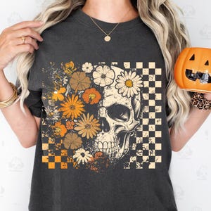 May include: Dark gray t-shirt featuring a skull design with floral and checkered patterns. The skull is adorned with daisies and other flowers in shades of orange, brown, and white. The shirt has a vintage, distressed look, perfect for Halloween or everyday wear.