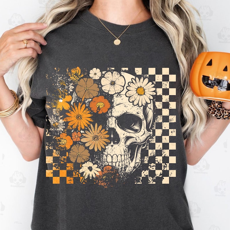 May include: Dark gray t-shirt featuring a skull design with floral and checkered patterns. The skull is adorned with daisies and other flowers in shades of orange, brown, and white. The shirt has a vintage, distressed look, perfect for Halloween or everyday wear.