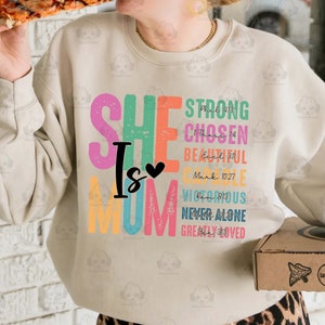 K&ouml;nnte beinhalten: Ein beiger Pullover mit den Worten "SHE IS MOM" in bunten Buchstaben. Die Buchstaben sind mit einem abgenutzten, Vintage-Look verziert. Jeder Buchstabe ist mit einem Bibelvers gepaart.