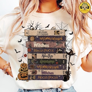 May include: A cream-colored t-shirt featuring a graphic of stacked VHS tapes labeled with movie titles including The Nightmare Before Christmas, Halloweentown, The Witches, Hocus Pocus, Addams Family, and Goosebumps, surrounded by spiderwebs and pumpkins.