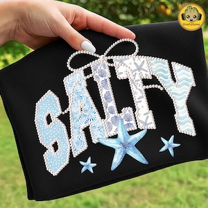 Puede incluir: Toalla de playa negra con la palabra "SALTY" en azul claro y blanco, decorada con conchas marinas y estrellas de mar. Las letras están delineadas con un borde perlado. La toalla se muestra sobre un fondo verde.