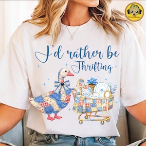 May include: White t-shirt with the text "I'd rather be Thrifting" in blue script. The graphic features a patchwork goose with a bow, and a shopping cart filled with dishes and a lamp. The design has a vintage, homespun aesthetic.