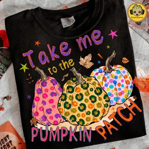 May include: A black t-shirt featuring three pumpkins with colourful patterns, including pink floral, green leopard print, and multicoloured polka dots. The text reads "Take me to the PUMPKIN PATCH" in purple and orange letters, accented by small stars and leaves.