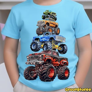 May include: A light blue t-shirt featuring a stack of five colorful monster trucks. The trucks are in shades of green, orange, blue, and red, with large tires and detailed graphics. The shirt has the text "GrootStoree" at the bottom.