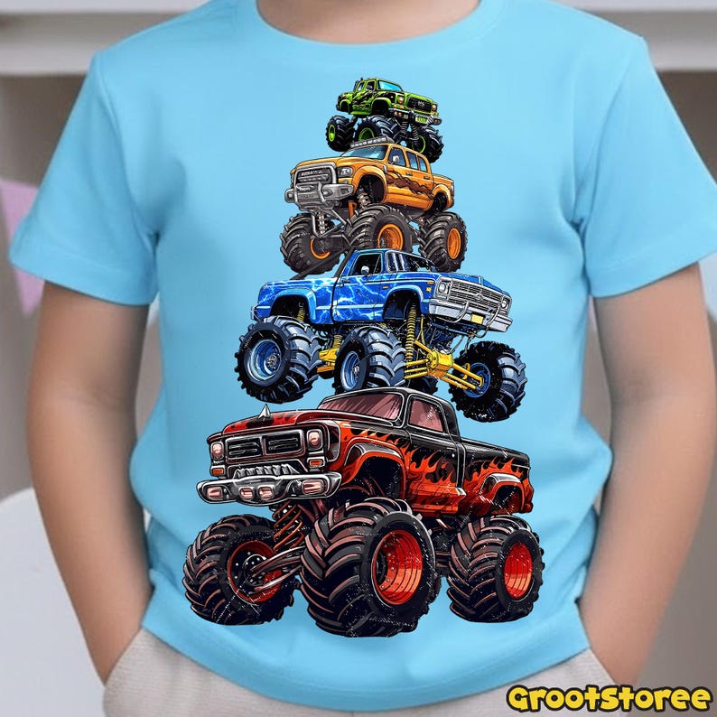 May include: A light blue t-shirt featuring a stack of five colorful monster trucks. The trucks are in shades of green, orange, blue, and red, with large tires and detailed graphics. The shirt has the text "GrootStoree" at the bottom.