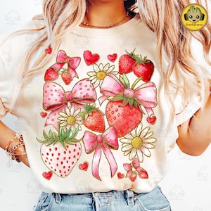 May include: A white t-shirt with a pink and red design featuring strawberries, bows, and flowers. The design is centered on the shirt and is surrounded by a white background.