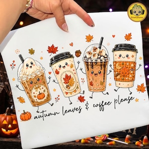 May include: A white t-shirt featuring a graphic of four anthropomorphic coffee cups with smiling faces, decorated with autumn leaves, pumpkins, and marshmallows. The text below reads, "autumn leaves & coffee please" in a black cursive script.