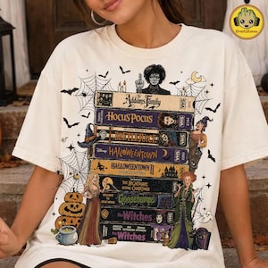 May include: A cream-colored t-shirt featuring a graphic print of a stack of VHS tapes labeled with movie titles including The Addams Family, Hocus Pocus, Beetlejuice, Halloweentown, The Nightmare Before Christmas, Goosebumps, The Witches, and The Witches.