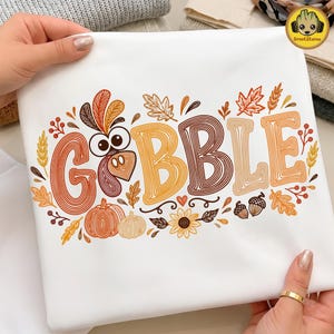 May include: A white t-shirt featuring a graphic print with the text GOBBLE in large, line-art style letters. The design includes a turkey head, pumpkins, autumn leaves, acorns, and a sunflower in shades of orange, brown, and yellow.