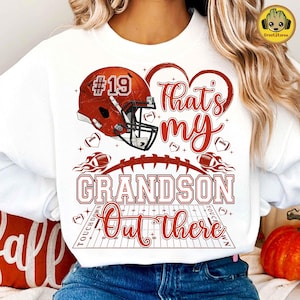 Peut inclure: Sweat-shirt blanc avec un casque de football américain rouge et le texte "That's my Grandson Out there." Le motif comprend des ballons de football, des cœurs et un terrain. Le numéro 19 est sur le casque.