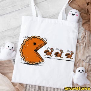 May include: White tote bag featuring a humorous Thanksgiving design. The graphic depicts a pie slice chasing three turkeys, reminiscent of a classic video game. The bag has two white handles and is made of a durable fabric.