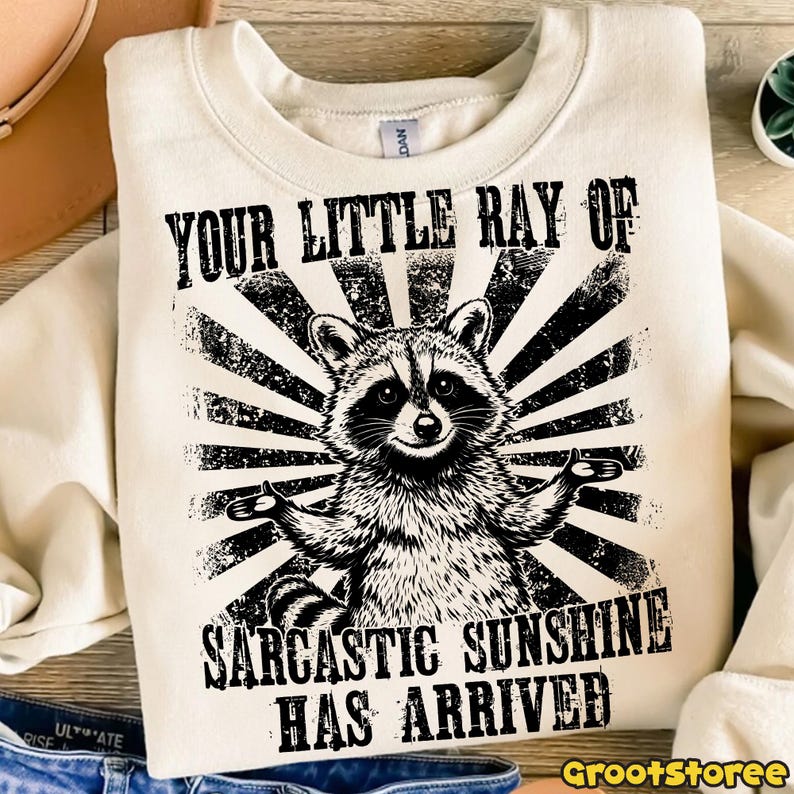 May include: Cream-coloured sweatshirt featuring a black and white raccoon graphic with outstretched arms, set against a sunburst design. The text reads "YOUR LITTLE RAY OF SARCASTIC SUNSHINE HAS ARRIVED."