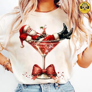May include: A cream-colored t-shirt featuring a festive graphic of Santa Claus relaxing in a large red martini glass. Santa is wearing his traditional red suit and hat, holding a cocktail. A red bow adorns the base of the glass, with sparkling accents.