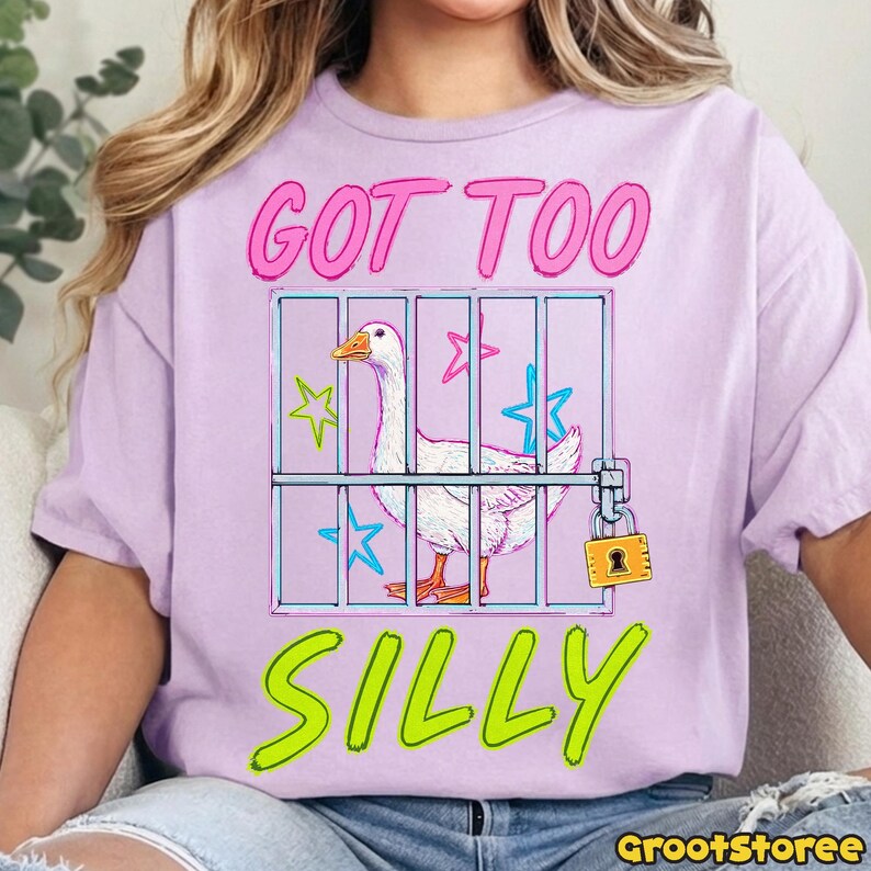 K&ouml;nnte beinhalten: Ein helllila T-Shirt mit den Worten "GOT TOO SILLY" in Pink und Gr&uuml;n. Die Grafik zeigt eine wei&szlig;e Gans in einem K&auml;fig mit Vorh&auml;ngeschloss, umgeben von Sternen. Ein l&auml;ssiges Kleidungsst&uuml;ck.