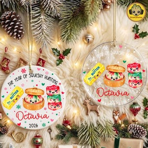 May include: Two round Christmas ornaments, one white ceramic and one clear acrylic, featuring the text THE YEAR OF SQUISHY HUNTING, Octavia, and 2026. Graphics include a yellow butter stick, a dumpling in a steamer, and three stacked squishy toys.