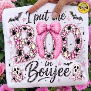 May include: A white t-shirt featuring the text I put the BOO in Boujee. The letters B and O are filled with pink, black, and clear rhinestones. A large pink fuzzy bow sits between the letters. The design includes black spiderwebs, bats, and ghosts.