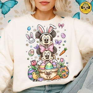 May include: Cream-colored sweatshirt featuring a colorful Easter-themed graphic. The design includes Mickey and Minnie Mouse dressed as bunnies, surrounded by Easter eggs, carrots, butterflies, and flowers. The graphic is detailed and vibrant.