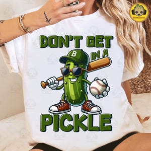 May include: White t-shirt with a green cartoon pickle wearing a baseball cap and sunglasses, holding a baseball bat and a baseball. The text on the shirt reads "Don't get in a pickle".