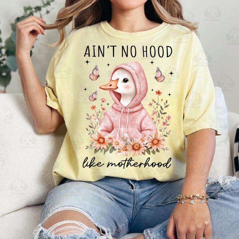 May include: A yellow t-shirt with a cartoon duck wearing a pink hoodie and surrounded by flowers. The text on the shirt reads "Ain't no hood like motherhood".