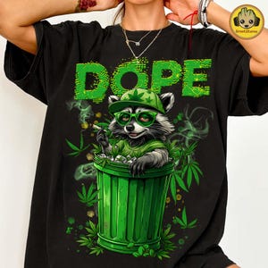 Puede incluir: Camiseta negra con un gráfico de un mapache con sombrero y gafas, fumando un cigarrillo y sentado en un cubo de basura verde. La palabra "DOPE" está impresa en verde encima del mapache.