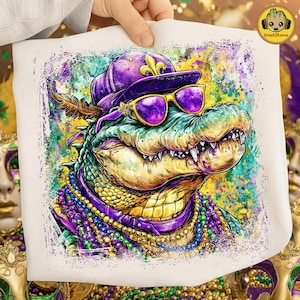 Könnte beinhalten: Weißes T-Shirt mit einer farbenfrohen Illustration eines Alligators mit lila Hut, Sonnenbrille und Mardi Gras-Perlen. Der Alligator ist detailliert mit grünen, gelben und lila Farbtönen. Der Hintergrund hat einen aquarellartigen Effekt.