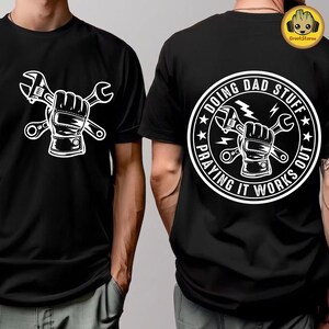 May include: A black t-shirt featuring a white graphic of a gloved hand holding two crossed wrenches on the front. The back displays a circular emblem with the same wrench graphic and the text "DOING DAD STUFF PRAYING IT WORKS OUT".