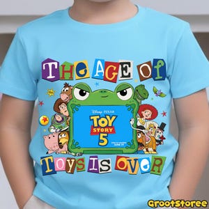 May include: A light blue short-sleeve t-shirt featuring a graphic print of various Toy Story characters surrounding a green frog-shaped frame. The text reads THE AGE OF TOY STORY 5 TOYS IS OVER, with Disney Pixar and June 19 printed inside.