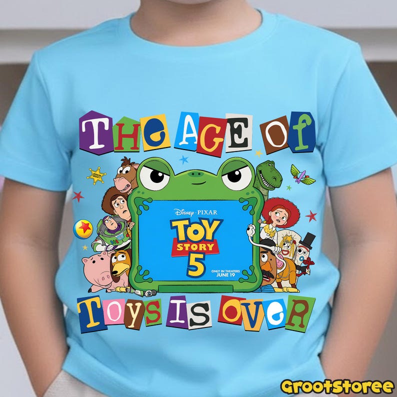 May include: A light blue short-sleeve t-shirt featuring a graphic print of various Toy Story characters surrounding a green frog-shaped frame. The text reads THE AGE OF TOY STORY 5 TOYS IS OVER, with Disney Pixar and June 19 printed inside.