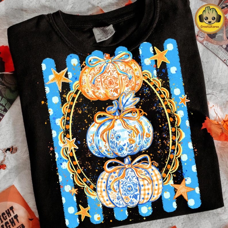 Puede incluir: Una camiseta negra con un gr&aacute;fico de tres calabazas apiladas dentro de un marco ovalado dorado ornamentado. Las calabazas est&aacute;n decoradas con patrones florales y de cuadros en azul y naranja con lazos. El fondo incluye rayas verticales azules.