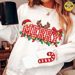 May include: A white long-sleeve sweatshirt featuring the red and white checkered text MERRY, decorated with a Santa hat, reindeer antlers, a red bow, and colorful Christmas lights. The sleeve displays a red and white candy cane with the name Octavia.