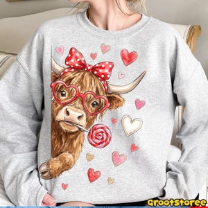 May include: A gray sweatshirt featuring a brown Highland cow wearing heart-shaped glasses and a red polka dot bow. The cow is holding a lollipop and surrounded by heart graphics. The text "GrootStoree" is at the bottom right.