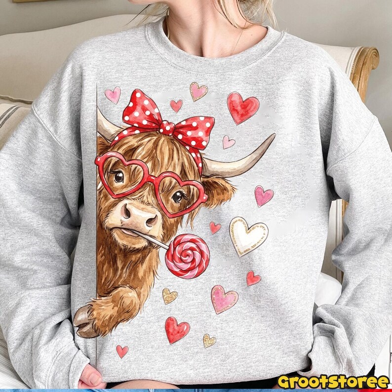 May include: A gray sweatshirt featuring a brown Highland cow wearing heart-shaped glasses and a red polka dot bow. The cow is holding a lollipop and surrounded by heart graphics. The text "GrootStoree" is at the bottom right.