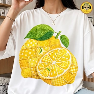 Puede incluir: Camiseta blanca con un diseño de limón grande en estilo patchwork. El limón está hecho de telas amarillas y blancas, con hojas verdes y un limón cortado que muestra el interior. El diseño está centrado en la camiseta.