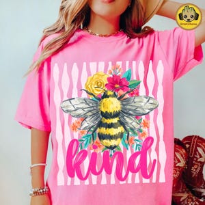 May include: Pink t-shirt featuring a large bee illustration with yellow and black stripes, surrounded by flowers. The word "kind" is written in pink script below the bee. The background has vertical white and pink stripes.