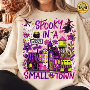 Può includere: Una felpa beige con una stampa grafica colorata che riporta il testo "SPOOKY IN A SMALL TOWN". Il design include un fantasma, una strega su una scopa, un calderone, una torre idrica e diversi edifici nelle tonalità viola, rosa e verde.